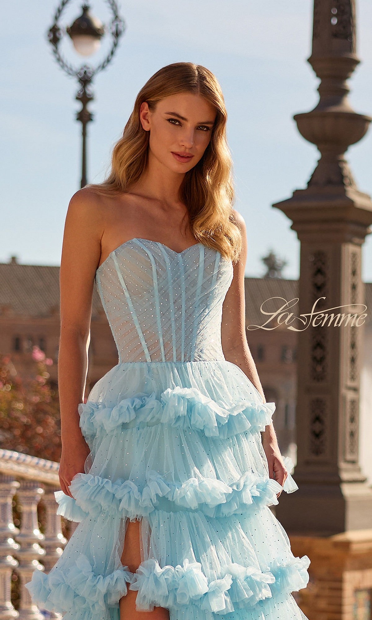 A woman stands outdoors wearing the La Femme 33812 long prom dress, featuring a strapless ruffled tulle bodice and layered skirt, with ornate streetlights and architectural details in the background.